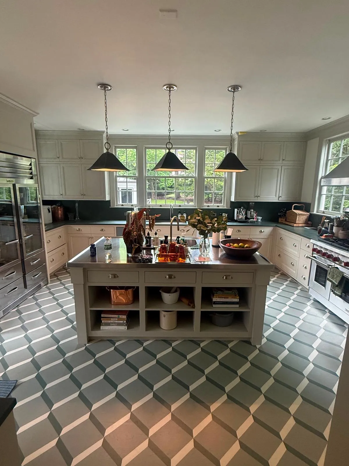 A finished kitchen with white cabinets, a gray island under three dark pendant lights, garden windows and a geometric gray and white patterned floor